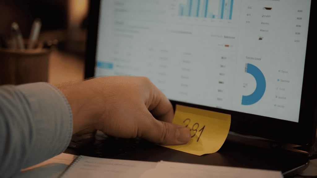A hand pressing a yellow sticky note with a single handwritten number onto the bottom edge of a laptop monitor, a softly blurred dashboard behind it